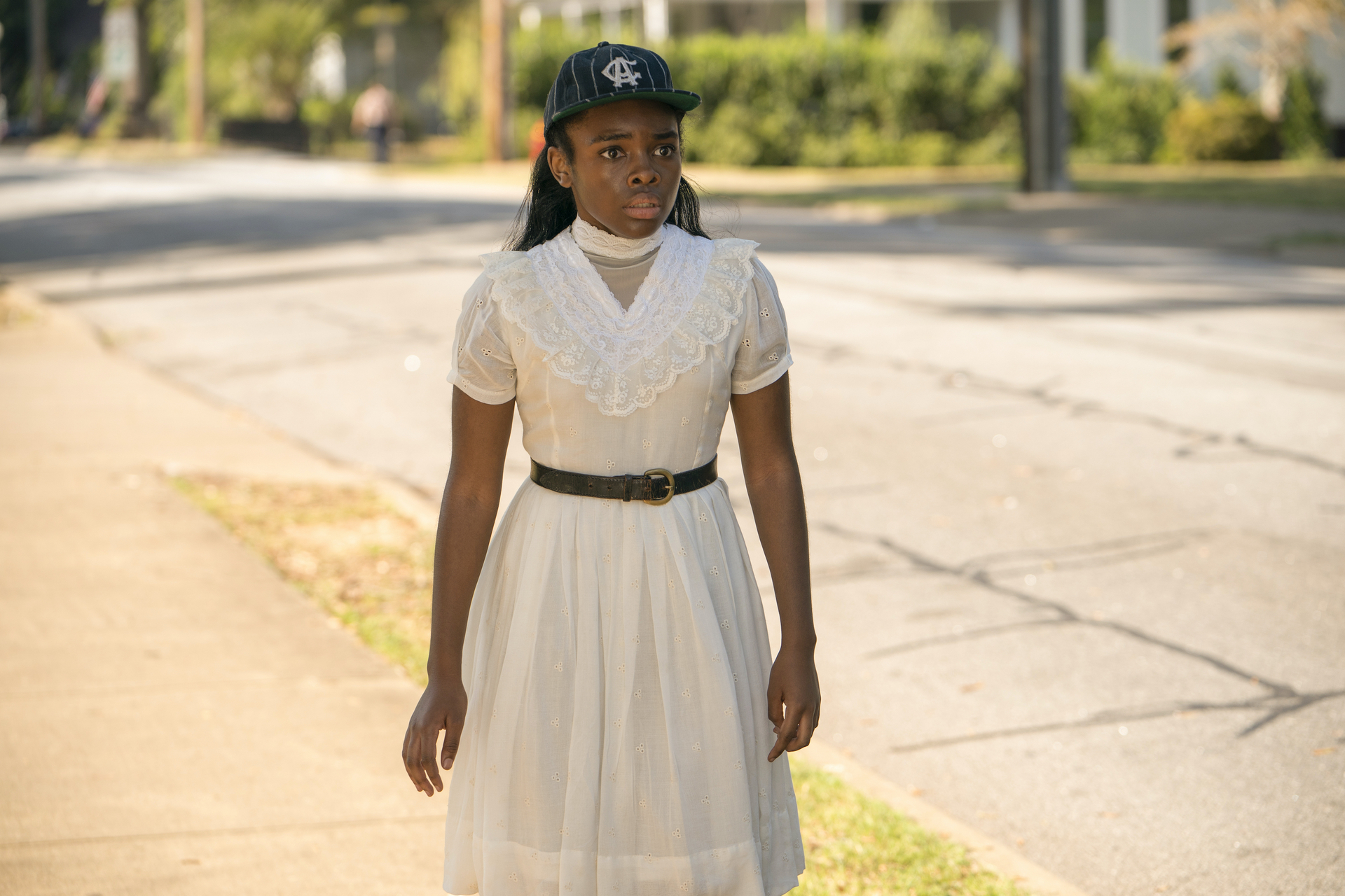 A young Black girl stands on a street wearing a white dress and baseball cap