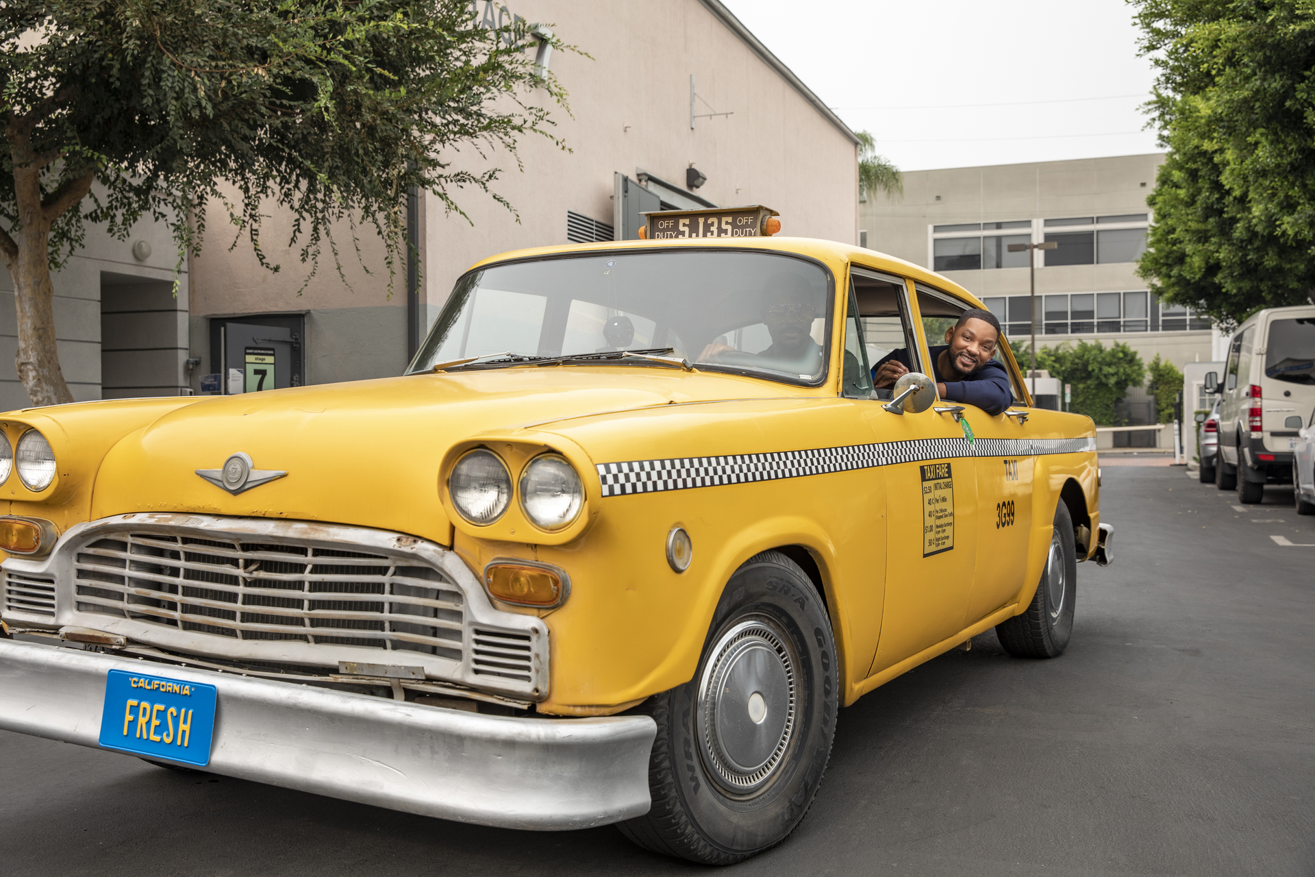 Will Smith sits in a yellow taxi with a blue license plate saying Fresh in yellow