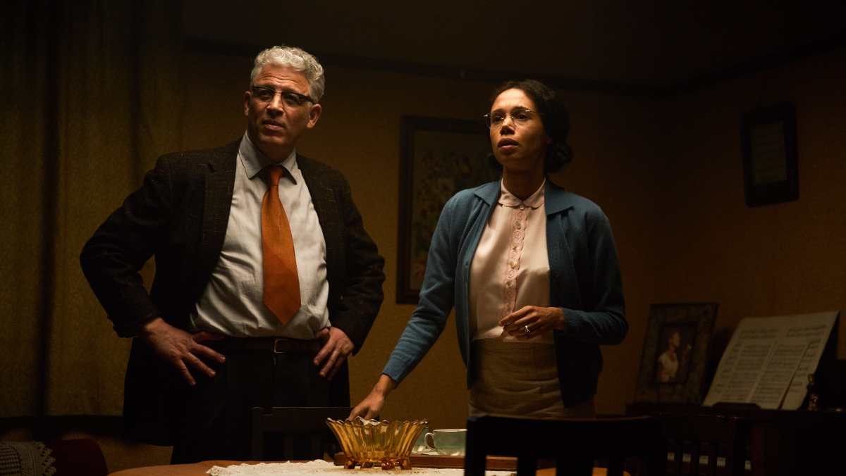 a woman depicting Rosa Parks stands beside a man wearing a dark suit in a dimly lit room 