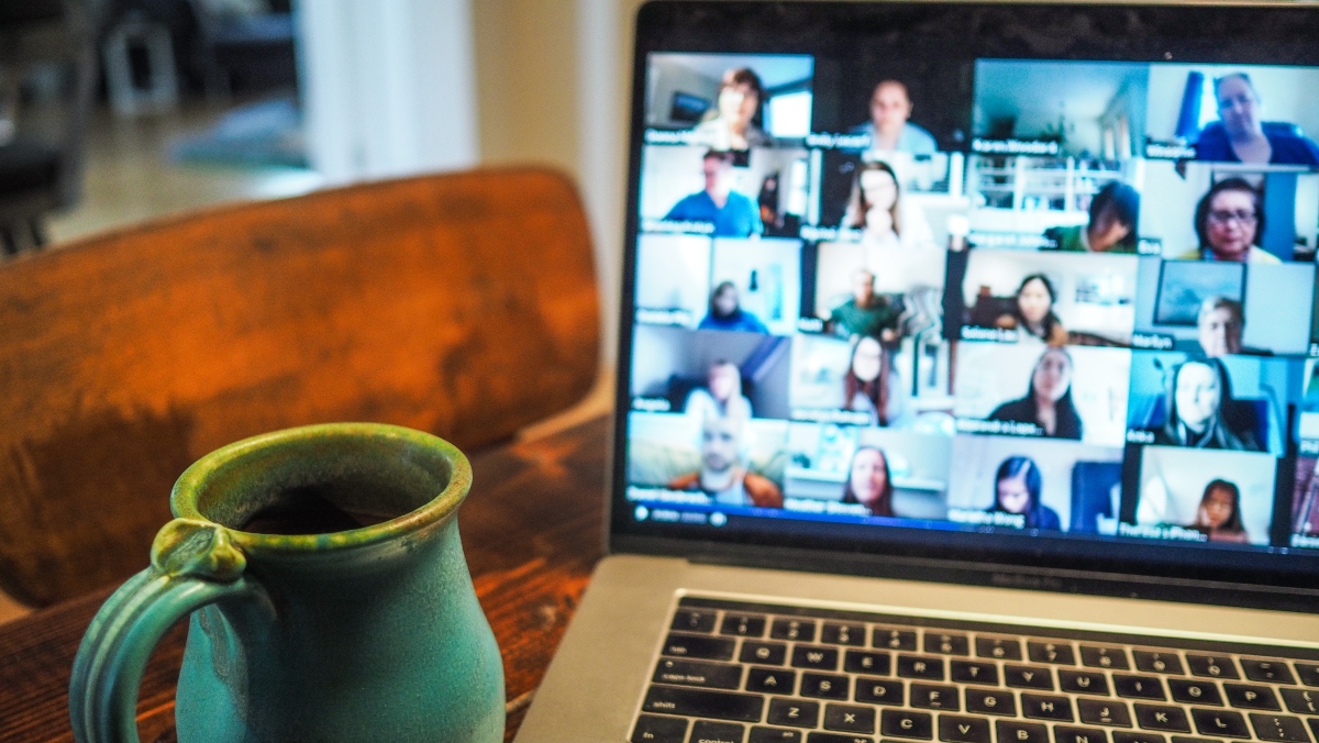 a stock image of a brown table with a turquiose coffee cup on the left side and a laptop with a Zoom call with several participants on the right