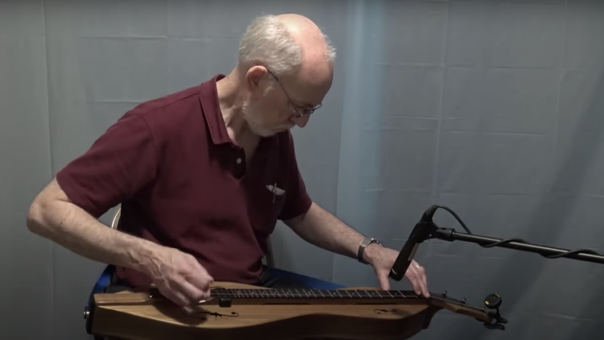 An older man plays a dulcimer against a gray background