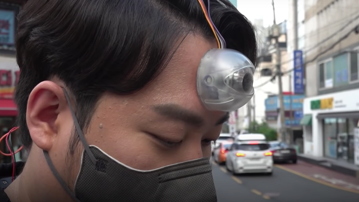 A man stands on the sidewalk in South Korea looking at his phone while also wearing a Third Eye camera.