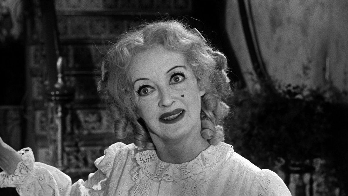 Baby Jane Hudson (Bette Davis) is an old woman wearing clothes and hair like that of a small child, smiles in a sinister manner with exaggerated pale face makeup and dark eye and lip makeup. She also has a small heart-shaped beauty mark on her left cheek. 