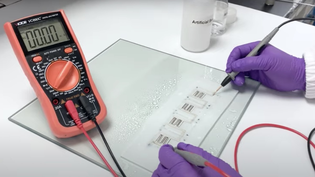 A voltage reader hooked up to a small board on a glass plate being worked on by someone wearing purple latex gloves