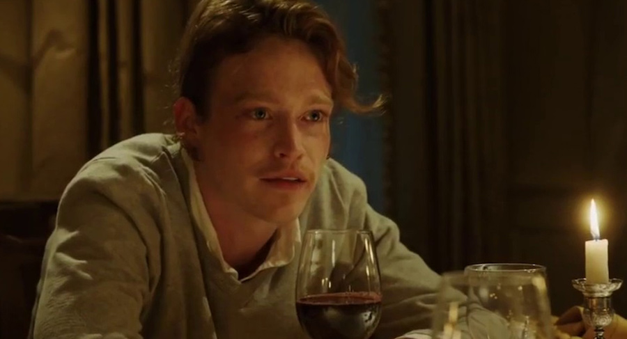 A young man with a terrible mustaches sits at a table with a glass of wine