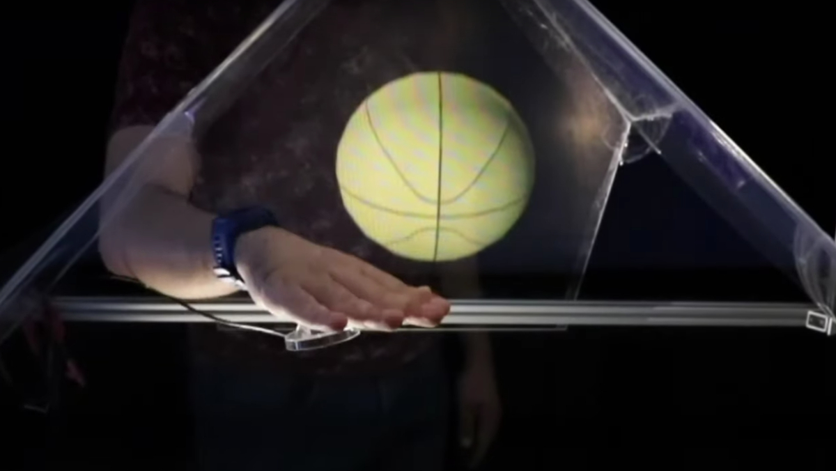 A man with his hand inside of a glass pyramid which contains a holographic basketball.