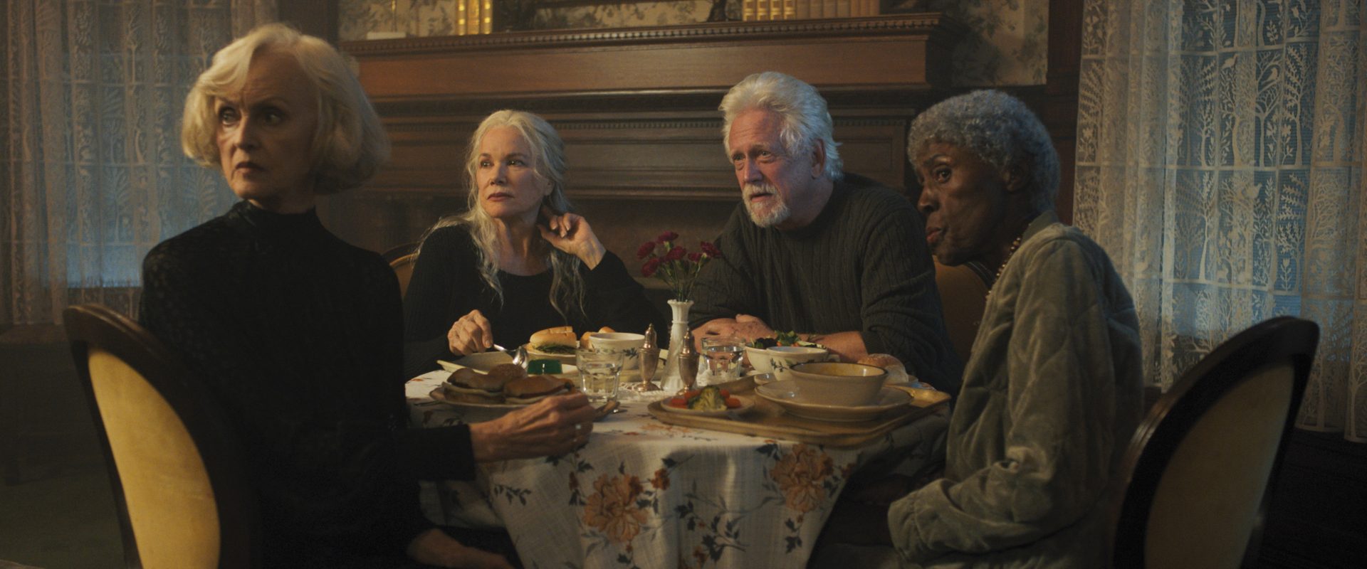 A still from The Manor shows a two older white women, an old white man, and an old Black woman sitting around a table looking worried