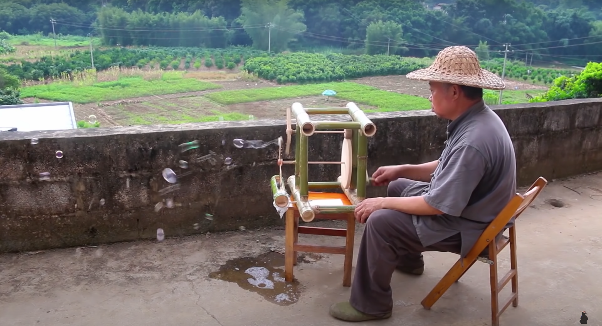 Master carpenter Grandpa Amu sits in a chair as he turns the wheel of a bamboo bubble machine spewing out bubbles.