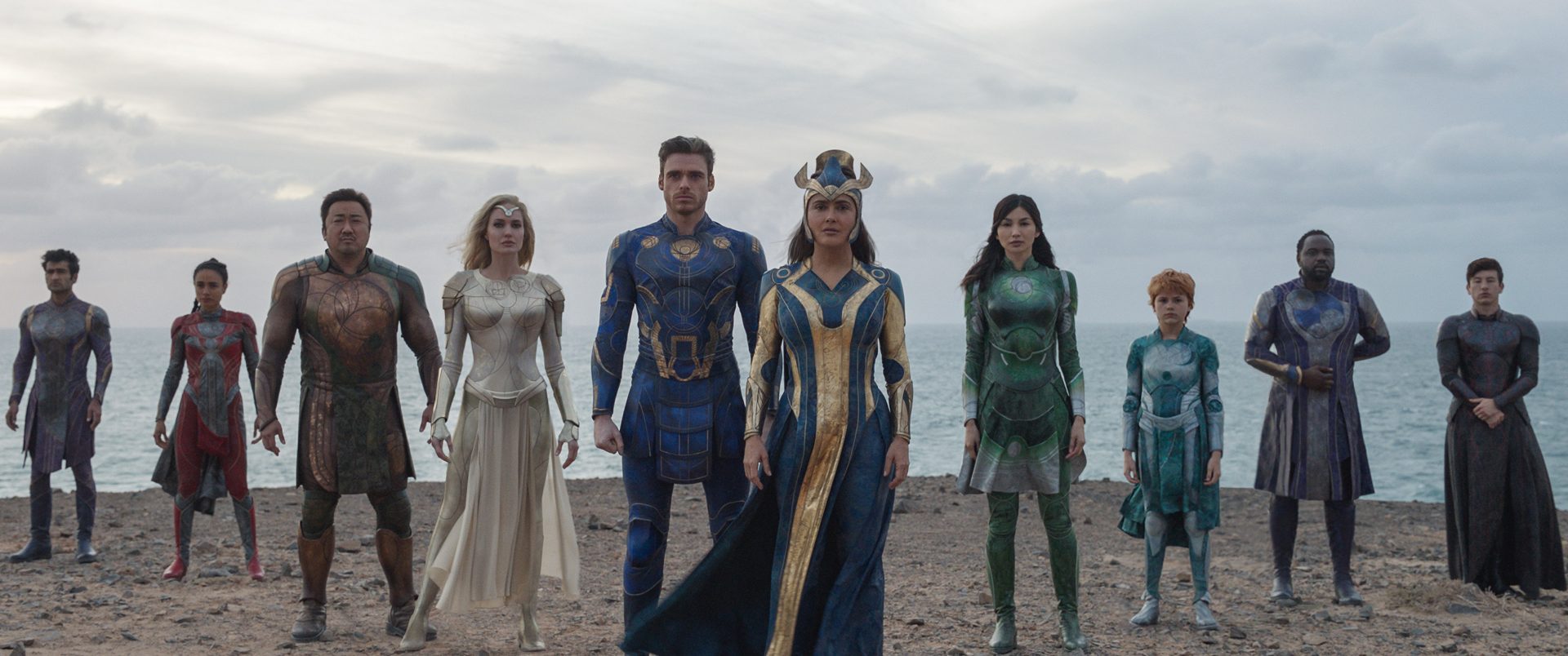 A still from Eternals shows the entire group standing on a beach in their supersuits