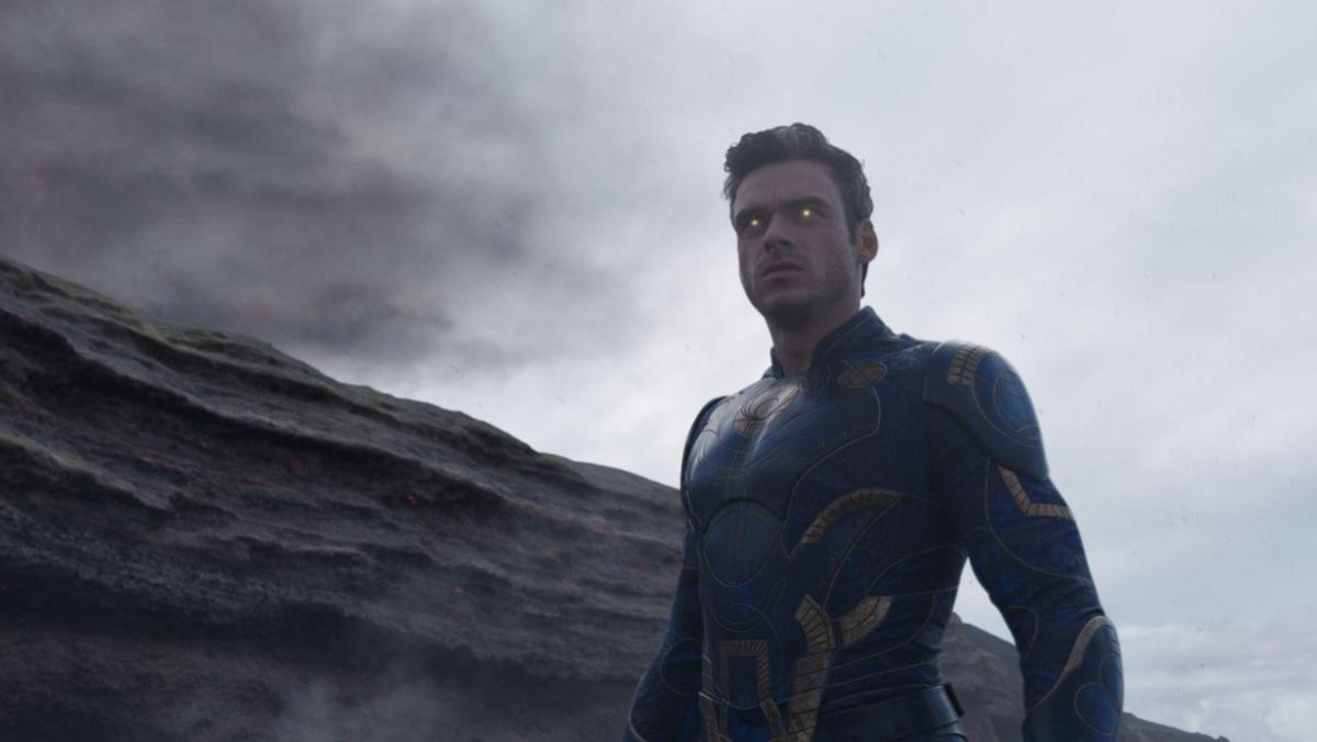 A still from Eternals shows Richard Madden as Ikaris glaring in front of a grey sky, his yellow eyes glow slightly