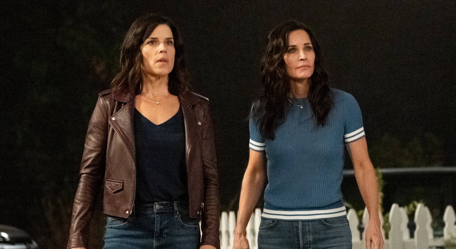 A still from Scream (2022) shows Neve Campbell and Courteney Cox as Sidney Prescott and Gale Weathers standing in front of a white picket fence film review.