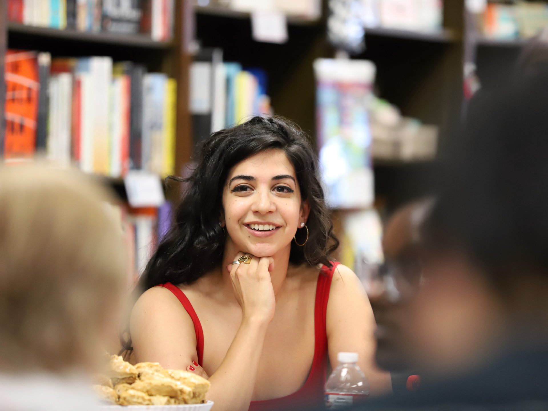 An image of the author Aminah Mae Safi shows a young woman with long brown hair her hand resting on her chin wearing a red vest top