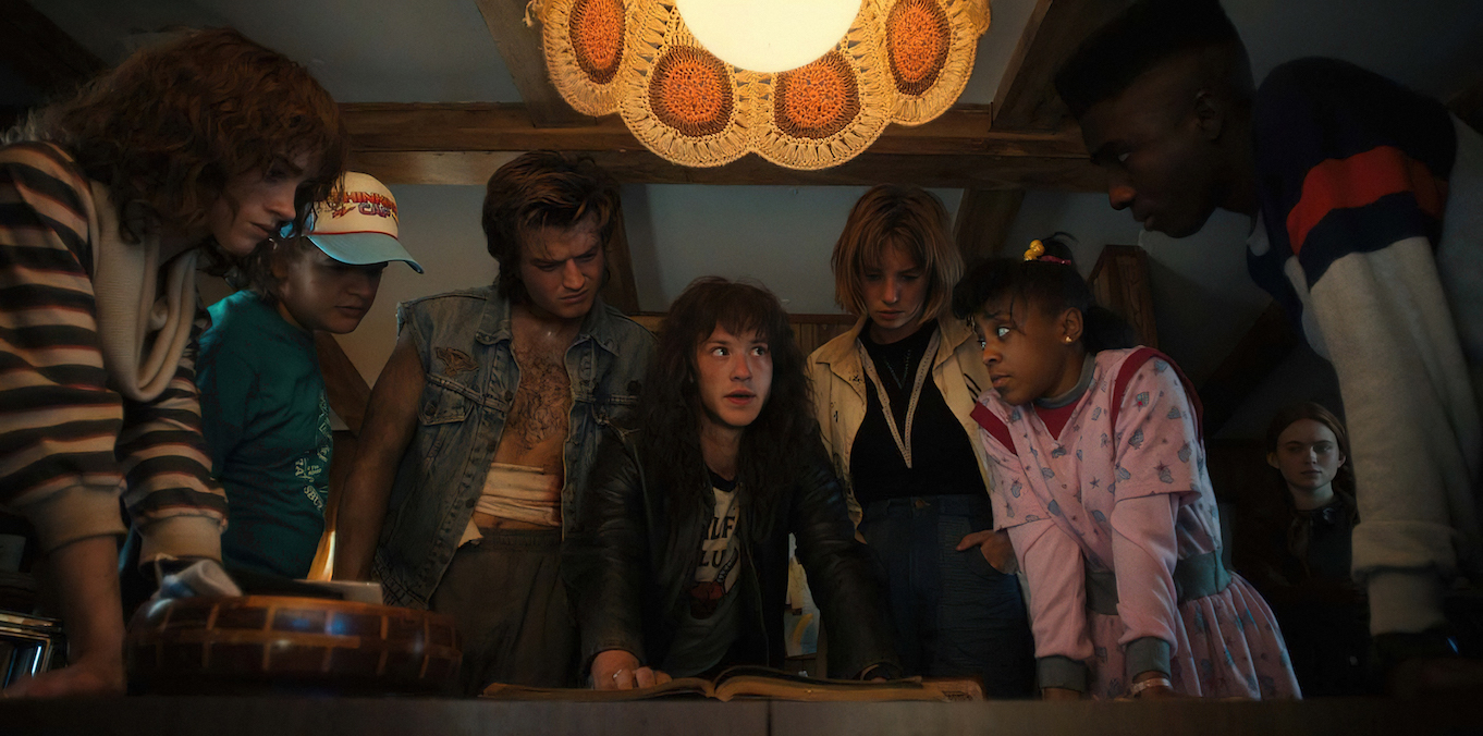 The Hawkins kids listen to Eddie while standing over a table on Stranger Things 4