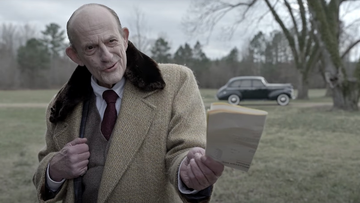 Christopher Lloyd in a winter coat holds out a book in Spirit Halloween - The Movie