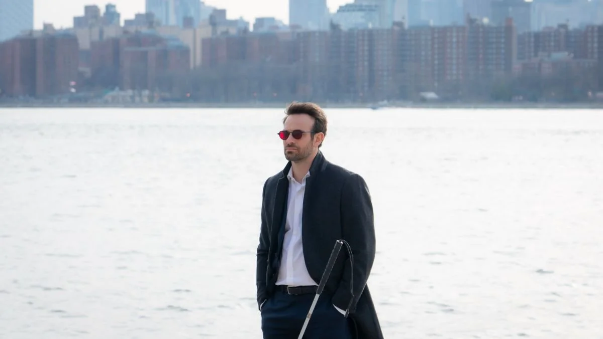 Charlie Cox, wearing a dark overcoat, white shirt, and red-tinted sunglasses stands by a waterfront with a city skyline in the background. He holds a white cane, indicating he is visually impaired. The water and city buildings are softly lit under a cloudy sky.