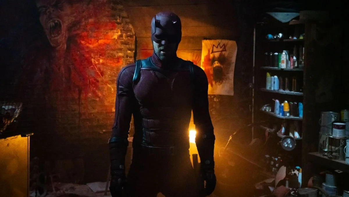 Daredevil, in a red armored superhero suit and mask stands in a dimly lit, cluttered room with exposed brick walls. The lighting casts dramatic shadows, emphasizing his muscular frame. Behind him, eerie artwork—including a screaming face and a blood-splattered canvas with a crown—adds to the ominous atmosphere. Shelves filled with spray paint cans and tools are visible on the right side of the image.