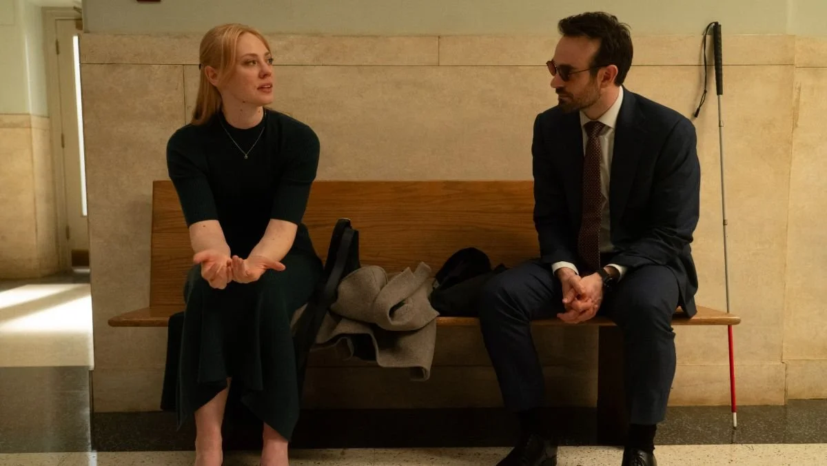 A woman and a man sit on a bench in a hallway talking. The man has a white cane beside him, indicating visual impairment. Coats are draped between them.