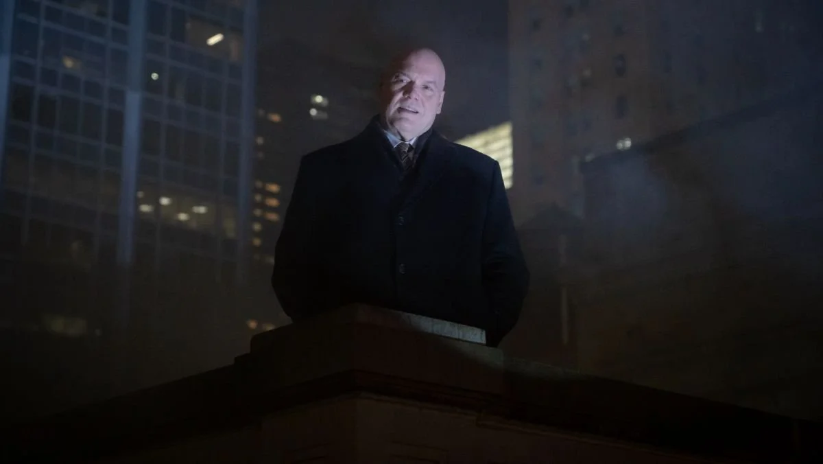A bald man in a dark coat stands on a rooftop at night, illuminated from below, with tall city buildings behind him in the mist.
