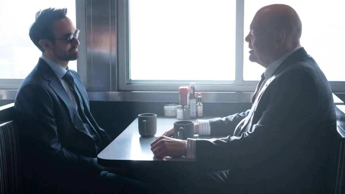 Two men in suits sit across from each other in a diner booth, talking over coffee with condiments on the table and bright light through the window.