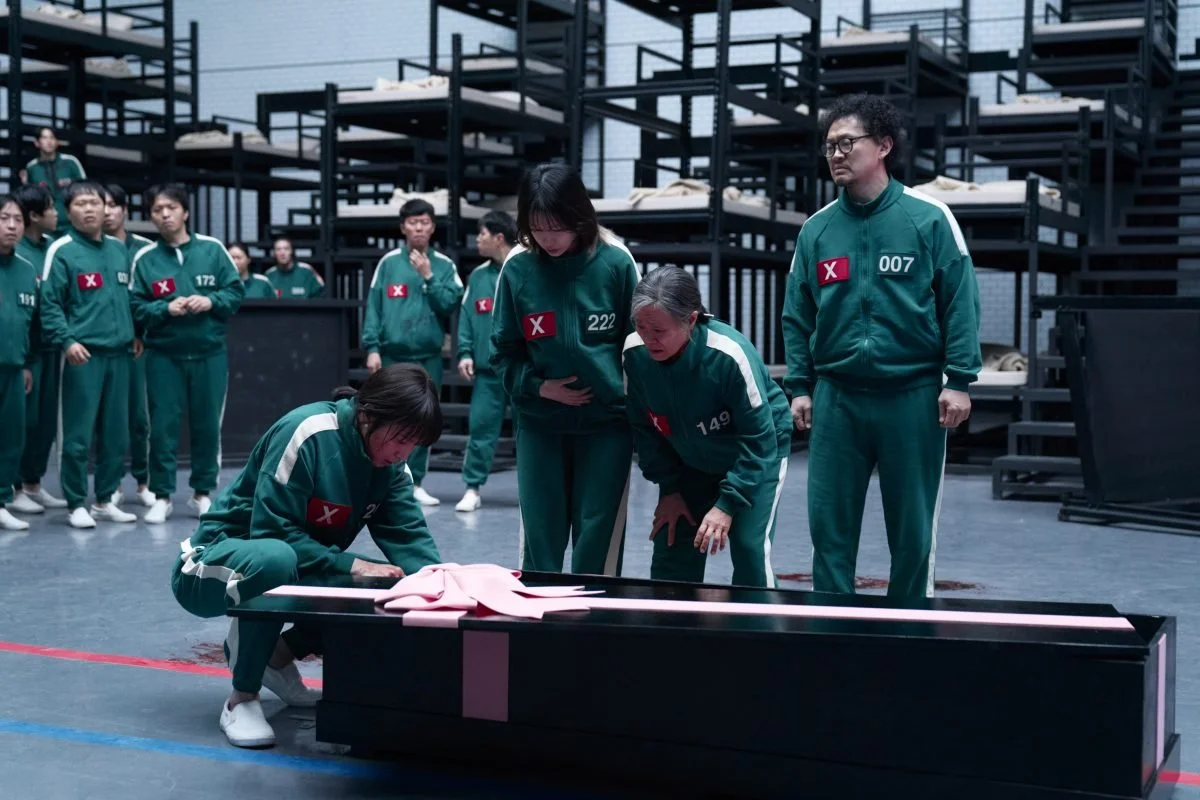 Players in green tracksuits around a black coffin with a pink bow on Squid Game
