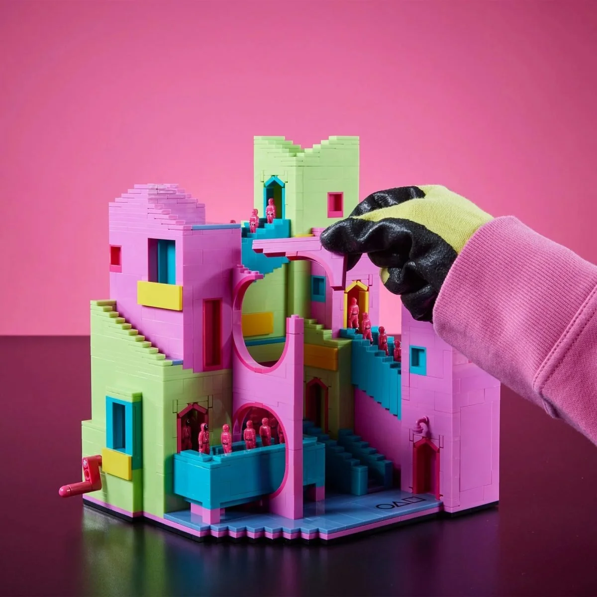 A gloved hand adjusts a piece of a pastel-colored Squid Game-themed building block set featuring pink, green, blue, and yellow stairs and walls, with small pink figurines positioned throughout the structure.