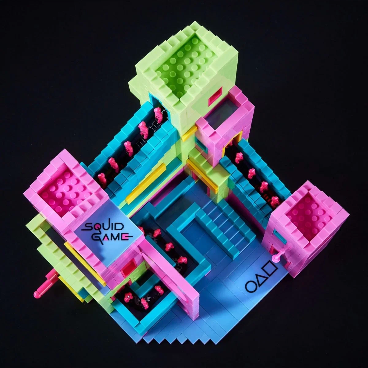Top-down view of a vibrant Squid Game-themed building block set featuring colorful staircases, pink guard figures, and labeled platforms on a black background.