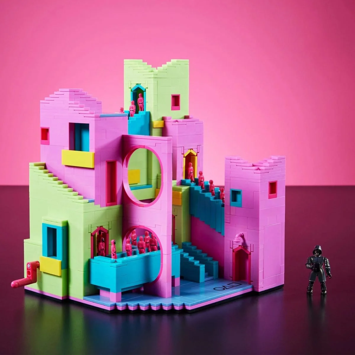 A colorful Squid Game-themed toy set made of building blocks with pastel pink, green, blue, and yellow walls, featuring multiple staircases and small pink figurines, alongside a black masked figure.