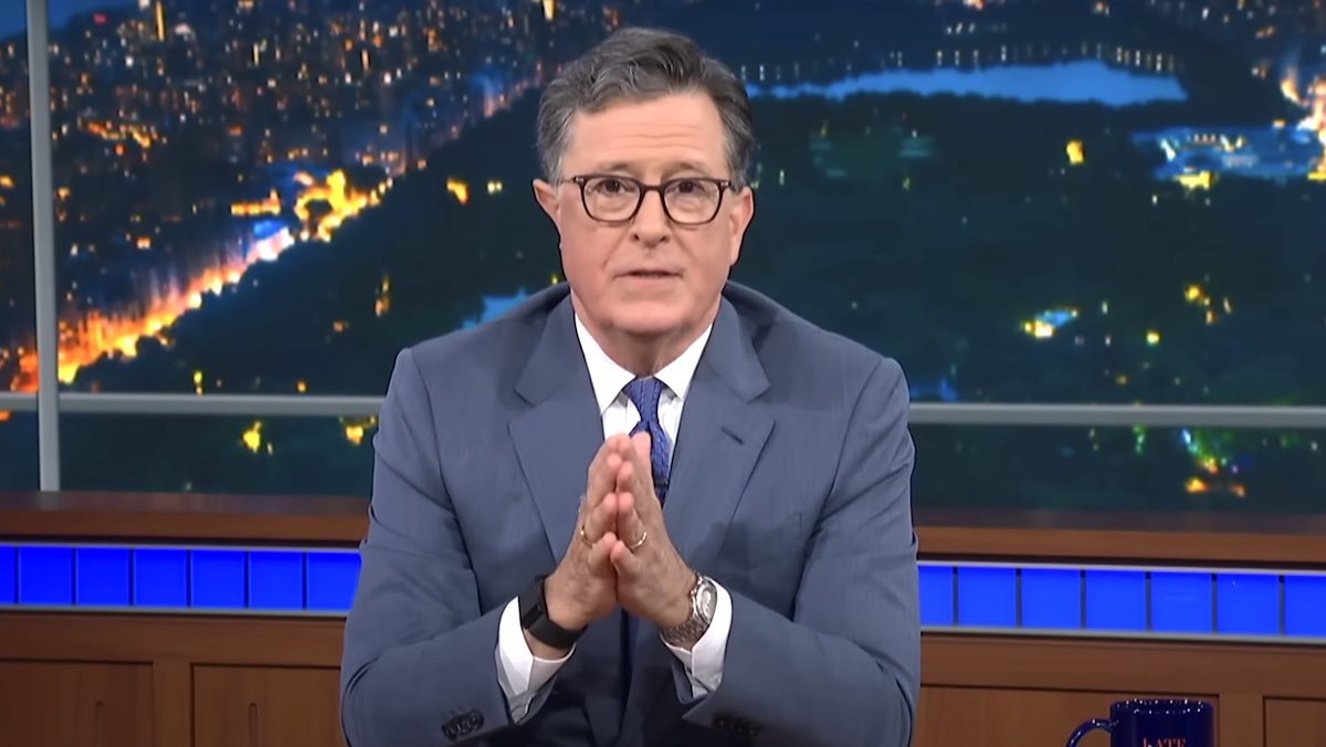 Stephen Colbert with his hand held together at his desk on The Late Show