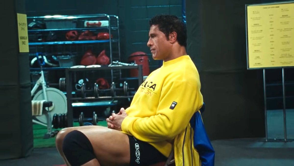 Dwayne Johnson with hair in a yellow sweatshirt sitting in profile in The Smashing Machine